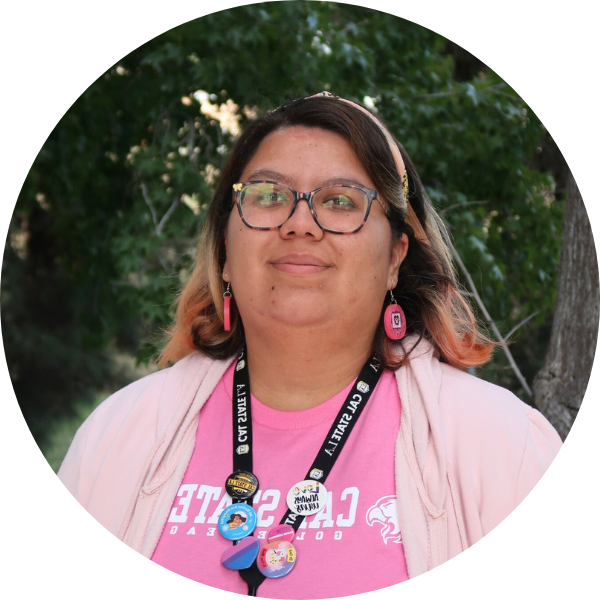 Woman in pink outfit and glasses smiling in front of foliage.